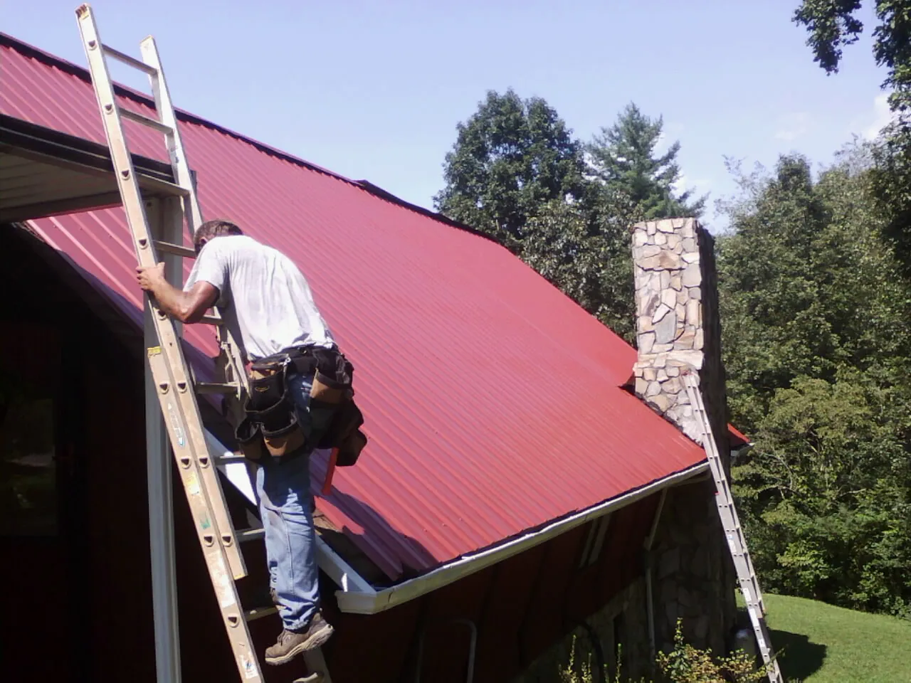 Roofer on a ladder beside a red metal roof and stone chimney — Soffit & Fascia Repair by Roofing Brothers in Flowood